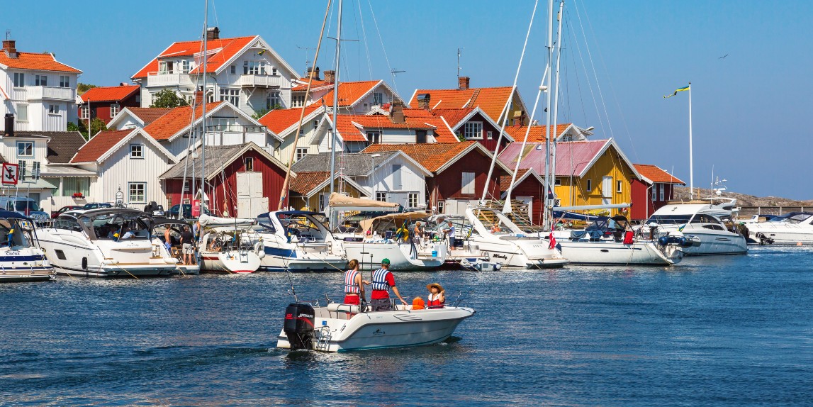 En familj är ute i båten vid en hamn bland många båtar, fina hus och svenska flaggor